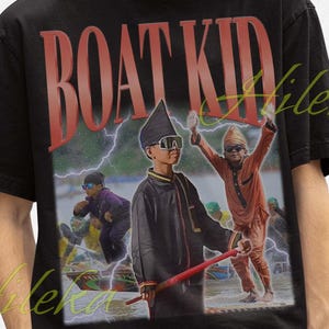 Op de afbeelding: Zwart T-shirt met de tekst "BOAT KID" in rode letters. Het shirt heeft een afbeelding van kinderen in kostuums met een bliksemachtergrond. De persoon op de foto draagt een zwarte vissershoed.