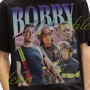 Può includere: T-shirt nero con grafica retrò di vigili del fuoco e la scritta "Bobby Nash". Il design include immagini di vigili del fuoco in uniforme. La persona nell'immagine indossa un cappello da pescatore nero.