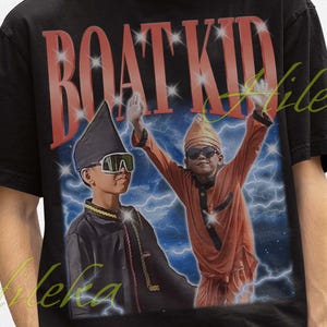 Op de afbeelding: Zwart T-shirt met een retro afbeelding van twee figuren met zonnebrillen en hoeden, en de tekst "BOAT KID" in het rood. De persoon op de afbeelding draagt een zwarte vissershoed. De afbeelding heeft een vintage stijl.