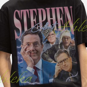 May include: Black t-shirt featuring a vintage-style graphic of Stephen Colbert with the name "STEPHEN" in a pink and white font. The design includes multiple images of Colbert. The person in the image is wearing a black bucket hat.