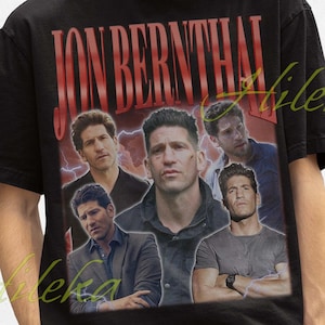 May include: Black t-shirt featuring a vintage-style graphic of Jon Bernthal with the text "JON BERNTHAL" in red. The design includes multiple images of the actor. The person in the image is wearing a black bucket hat.
