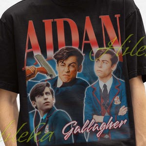 May include: Black t-shirt featuring a vintage-style graphic of Aidan Gallagher. The design includes the name "AIDAN" in large red letters above multiple images of the actor. The shirt is worn by a person wearing a black bucket hat.