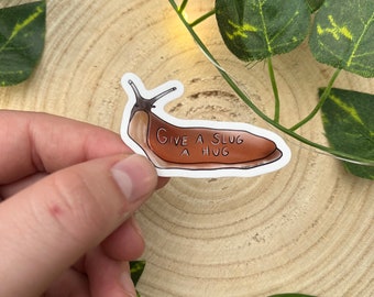 May include: A sticker featuring a brown slug with the text "GIVE A SLUG A HUG" in a handwritten font. The slug has a light brown body and a darker brown head. The sticker is held against a wooden background with green leaves.