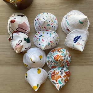 Puede incluir: Una colección de discos de lactancia reutilizables con varios diseños, incluyendo motivos florales, de hojas y de zorros. Los discos son blancos con detalles coloridos y están dispuestos sobre una superficie de madera. Una vela en un tarro de cristal marrón está en el fondo.