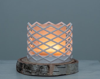 May include: A white, lattice-patterned candle holder with a lit candle inside, resting on a round, natural wood slice. The candle emits a warm, inviting glow. The holder's design creates a diamond pattern. The wood slice base adds a rustic touch.