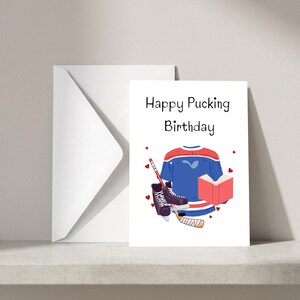 May include: A white greeting card with the text Happy Pucking Birthday in black font. The illustration features a blue, red, and white hockey jersey, black ice skates, a wooden hockey stick, and an open red book surrounded by small red hearts.