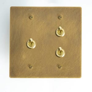 Peut inclure: Une plaque d'interrupteur carrée de couleur laiton avec trois interrupteurs à bascule et quatre vis. La plaque a une surface texturée et une finition métallique chaude. Les interrupteurs à bascule ont une extrémité arrondie en forme de boule.