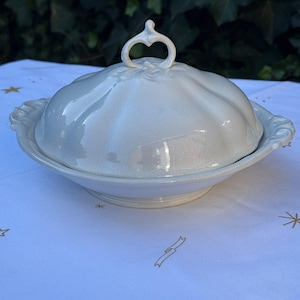 May include: A white ceramic serving dish with a matching lid, featuring a scalloped, pumpkin-like shape and a decorative heart-shaped handle on top. The dish has two side handles with embossed detailing and a glossy, crackled glaze finish.