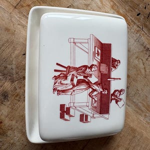 May include: Cream-colored rectangular ceramic napkin holder with a red illustration of figures and a printing press. The holder has a curved, raised edge on one side.