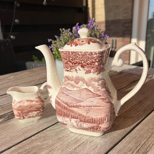 Puede incluir: Un juego de té de cerámica color crema con un diseño de paisaje en tonos rojos. El juego incluye una tetera con tapa y una jarrita para la crema. La tetera tiene un asa y un pico curvos. La jarrita tiene un asa y un pico pequeños.