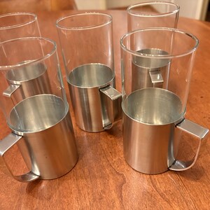 Könnte beinhalten: Sechs klare Glasbecher mit Edelstahl-Becherhaltern und Griffen sind auf einer Holzoberfläche angeordnet. Die Gläser haben eine zylindrische Form und die Metallhalterungen haben ein gebürstetes Finish.