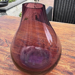 Op de afbeelding: Een doorschijnende, peervormige vaas van paars glas met een brede, ronde basis en een nauwere opening aan de bovenkant. Een ronde sticker op de bovenkant van het glas bevat de tekst "HAND MADE QUALITY" rondom een klein grafisch icoon.