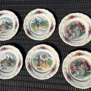 Puede incluir: Un juego de seis platos de cerámica blanca con bordes festoneados, cada uno con una ilustración colorida única de edificios o figuras en un paisaje, acentuado por motivos florales en rojo, azul y verde a lo largo de los bordes.