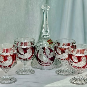 Op de afbeelding: Een karaf van helder en robijnrood glas met een bijpassende set van vier glazen op voet. Elk stuk is voorzien van geëtste ontwerpen van vogels op takken met bladeren. De karaf heeft een hoge, slanke hals en een ronde voet met vogelmotief.