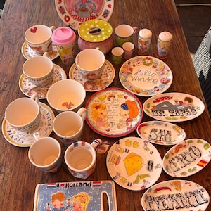 Puede incluir: Una colección de vajilla de cerámica que incluye varias tazas, platillos y platos con coloridas ilustraciones pintadas a mano de personas, comida y textos como I Love Holland, Hello Winter, Mr. Tapas, Sal, Frio y Picante sobre una superficie.