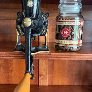 Op de afbeelding: Een zwarte gietijzeren koffiemolen met gouden accenten, voorzien van de tekst "SPONG & CO ENGLAND" en "D-E", naast een glazen pot gevuld met koffiebonen, met het etiket "DOUWE EGBERTS KOFFIE" en een rood-wit logo.