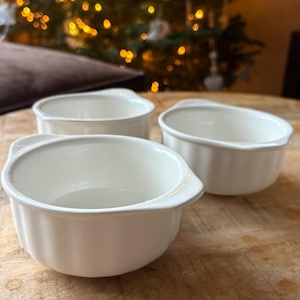 Può includere: Tre ciotole in ceramica bianco sporco con piccole maniglie su una superficie di legno. Le ciotole sono rotonde con un bordo leggermente svasato. Lo sfondo include un albero di Natale sfocato con luci calde e dorate.