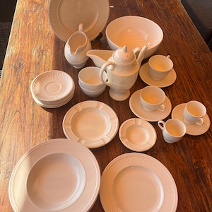 Puede incluir: Un conjunto de vajilla de cerámica color crema que incluye platos llanos, cuencos para sopa, platillos, tazas de té, una tetera, una salsera y una lechera. Cada pieza presenta una textura sutilmente acanalada en los bordes y un acabado liso.
