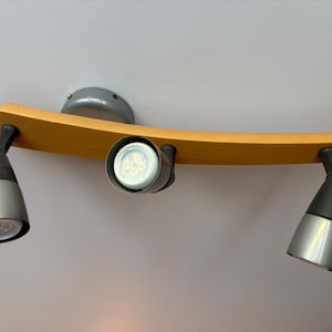 Könnte beinhalten: Eine Deckenleuchte mit einer geschwungenen, hellen Holzmontageschiene und drei verstellbaren Strahlern. Jeder Strahler verfügt über einen mattschwarzen Arm und einen konischen, silberfarbenen Metallschirm mit einer runden LED-Lampe.