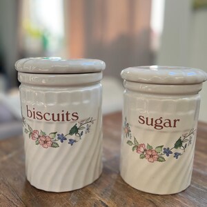 Puede incluir: Dos botes de cerámica blanca con diseños florales y tapas. El bote de la izquierda dice "galletas" y el bote de la derecha dice "azúcar".