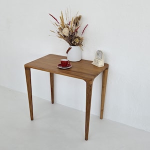 Puede incluir: Pequeña mesa rectangular de madera con cuatro patas delgadas. Una taza y un platillo rojos están sobre la mesa, junto a un jarrón blanco con flores secas y un pequeño reloj vintage. La mesa es de madera clara.