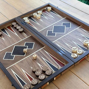May include: A wooden backgammon board featuring a dark walnut frame with inlaid geometric diamond patterns in black and white. The playing surface includes alternating black and red triangular points, accompanied by circular wooden checkers and two dice.
