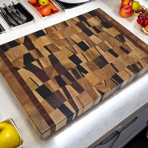 May include: A large, rectangular cutting board made of various types of wood. The board is made up of many small, rectangular pieces of wood, each with a different grain pattern. The board has a natural wood finish and is surrounded by a dark brown border.