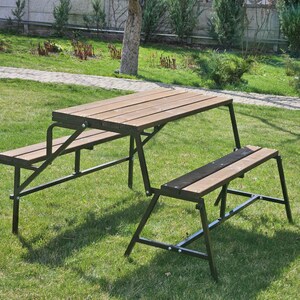 Könnte beinhalten: Ein Holzpicknicktisch mit zwei Bänken, beide aus Holz und Metall. Tisch und Bänke sind schwarz und haben ein rustikales Aussehen. Der Tisch ist zusammengeklappt und die Bänke sind heruntergeklappt.
