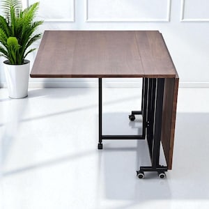 Könnte beinhalten: Ein brauner, hölzerner Klapptisch mit einem schwarzen Metallrahmen und Rollen. Der Tisch ist teilweise zusammengeklappt und gibt die Unterseite frei. Eine grüne Topfpflanze ist im Hintergrund zu sehen.