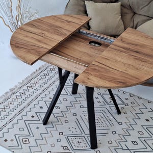 May include: A wooden, extendable dining table with a natural wood grain finish. The table has a black metal base and is partially extended, revealing the inner mechanism. It sits on a patterned rug.