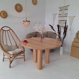 Könnte beinhalten: Ein runder Esstisch aus hellem Holz mit drei zylindrischen Beinen. Links steht ein Stuhl aus geflochtenem Rattan mit einem Kissen. Der Raum ist mit Hüten, Makramee-Wandbehängen und einer Pendelleuchte dekoriert. Mehrere verpackte Kartons sind an der Wand gestapelt.