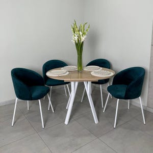 May include: A round dining table with a light wood top and white legs, surrounded by four teal velvet chairs. The table is set with four plates and a vase of white flowers.