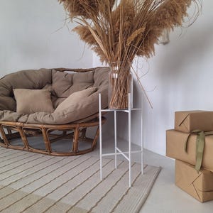May include: A brown wicker chair with two brown cushions and a white metal stand with a vase of dried pampas grass. There are three brown wrapped boxes on the floor.