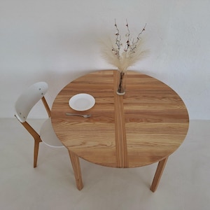 May include: A round wooden table with a light brown finish, a white plate, and a fork. A white chair with a wooden frame is positioned next to the table. A vase with decorative dried plants sits on the table.
