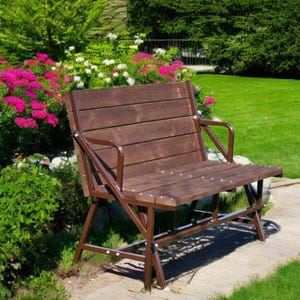 Può includere: Una panchina di legno marrone con una struttura in metallo si trova su un sentiero in un giardino. La panchina ha uno schienale e una seduta a doghe. Il giardino è circondato da erba verde e cespugli con fiori rosa.