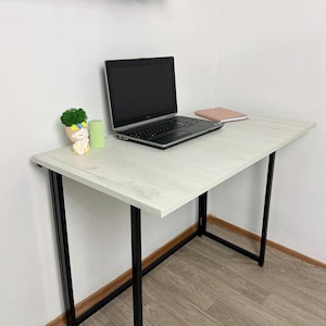 May include: A white and black desk with a laptop, notebook, and small decorative items. The desk has a light wood grain finish and black metal legs. A black laptop is open on the desk, with a pink notebook and a small decorative plant.