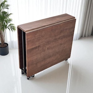 Peut inclure: Une table pliante en bois marron avec une bordure de couleur claire. La table est sur roulettes et semble être en position fermée. Une plante en pot est visible en arrière-plan, près d'un rideau blanc.