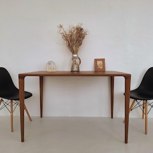 May include: A wooden rectangular table with four slender legs, flanked by two black chairs with wooden legs. A vase of dried flowers, a small framed picture, and a wooden calendar sit on the table. The scene is set against a white wall.