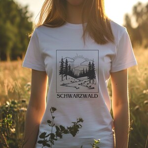 Ein weibliches Modell steht mitten in einer hohen Wiese. Sie trägt ein weisses T-Shirt mit Naturmotiv darauf. Auf dem Motiv ist der Schwarzwald, ein Bach, die Sonne und kleine Hügel abgebildet. Das Motiv ist schwarz, eckig und Minimalistisch.