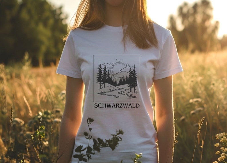 Ein weibliches Modell steht mitten in einer hohen Wiese. Sie trägt ein weisses T-Shirt mit Naturmotiv darauf. Auf dem Motiv ist der Schwarzwald, ein Bach, die Sonne und kleine Hügel abgebildet. Das Motiv ist schwarz, eckig und Minimalistisch.