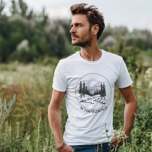 Ein männliches Modell steht mitten in einer grünen Wiese. Er trägt ein weisses T-Shirt mit Naturmotiv darauf. Auf dem Motiv ist der Schwarzwald, ein Bach, die Sonne und kleine Hügel abgebildet. Das Motiv ist schwarz, eckig und Minimalistisch.