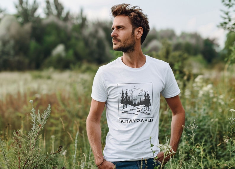 Ein männliches Modell steht mitten in einer grünen Wiese. Er trägt ein weisses T-Shirt mit Naturmotiv darauf. Auf dem Motiv ist der Schwarzwald, ein Bach, die Sonne und kleine Hügel abgebildet. Das Motiv ist schwarz, eckig und Minimalistisch.