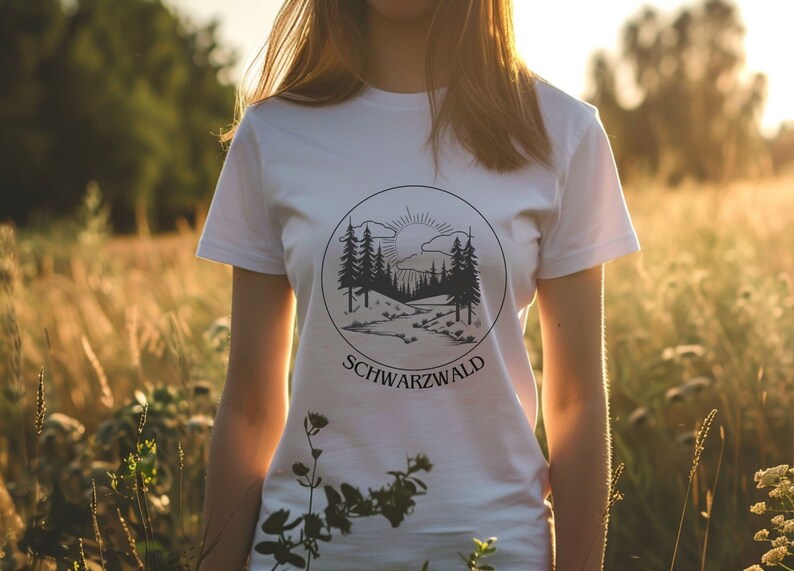 Ein weibliches Modell steht mitten in einer hohen Wiese. Sie trägt ein weisses T-Shirt mit Naturmotiv darauf. Auf dem Motiv ist der Schwarzwald, ein Bach, die Sonne und kleine Hügel abgebildet. Das Motiv ist schwarz, rund und Minimalistisch.