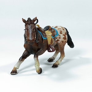 May include: A brown and white spotted toy horse with a brown saddle and blue blanket. The horse is running with its head turned to the side.