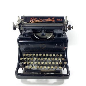Può includere: Una macchina da scrivere Rheinmetall Mod. 9 nera. La tastiera ha tasti rotondi e una leva di ritorno carrello. Il nome "Rheinmetall" è visualizzato in lettere rosse e dorate sopra i tasti.