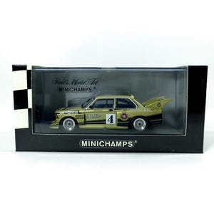 May include: A gold 1:18 scale model car of a BMW 3 Series race car with the number 4 on the side. The car is on a black display base with the Minichamps logo. The car is a part of the Minichamps World of  Model Art collection.