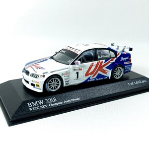 May include: A white, blue, and red BMW 320i model car. The car features the UK flag design and the text "BMW Team UK". The model is on a black base with the text "BMW 320i WTCC 2005 Champion: Andy Priaulx".