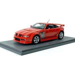 Op de afbeelding: Een rode model sportwagen met een spoiler en zilveren wielen staat op een grijs platform. De miniatuurauto heeft gedetailleerde kenmerken zoals een grille, zijopeningen en een kleine antenne.
