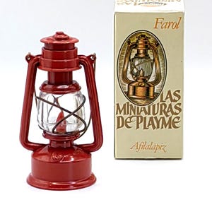 May include: A miniature red lantern with a clear glass globe and a handle, next to a box with the word "Farol" and an illustration of a lantern. The lantern is a decorative item.
