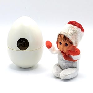 May include: A white egg-shaped container with a gold emblem and a small doll wearing a white and red outfit. The doll has blue eyes and is seated. The egg-shaped container is next to the doll.